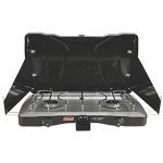 Portable Coleman Triton 2-Burner Propane Stove with wind-blocking panels and dual burners, set up in a picturesque outdoor setting.